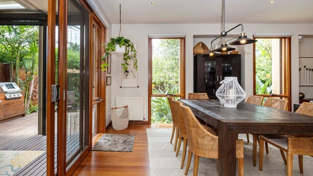 A dining area with a wooden table and wicker chairs, large windows with a view of greenery, and a hanging light fixture. - Pet Friendly - Bayside Oasis in Highett (Highett)