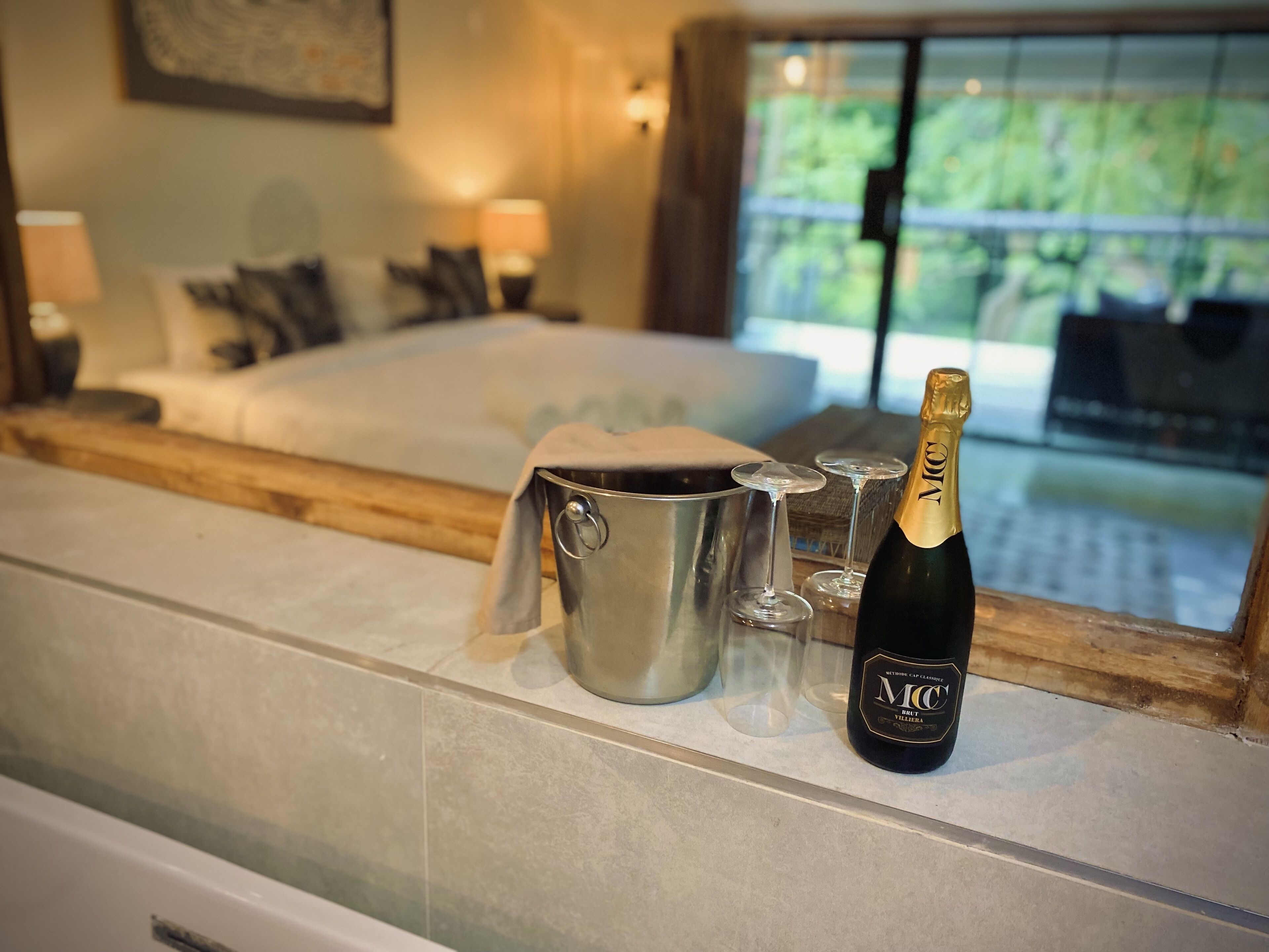 A bedroom with a bed, bedside lamps, a minibar with a bottle of champagne, and a view of greenery through a window.
