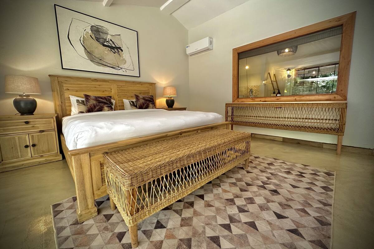 Ein Schlafzimmer mit einem großen Bett, einem Holz-Kopfteil, einer Korbmöbelbank, einem gemusterten Teppich und einem großen Spiegel.