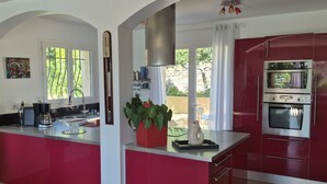 A kitchen with red cabinets, a black stove, and a view of the outdoors. - Pretty villa with sea view and pool **** (Bormes-les-Mimosas)