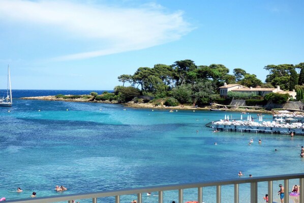 A coastal view with clear blue waters, a sailboat, and a beach area with people and umbrellas.
