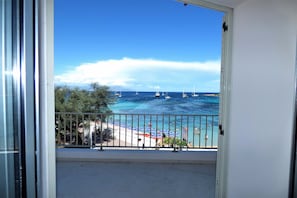 Uma varanda com vista clara para o mar, uma praia com guarda-sóis e veleiros.