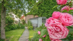 A garden pathway leading to a shed, surrounded by lush greenery and pink roses. - Vacation home Hus rode Dör for up to 5 people (Oldenswort)