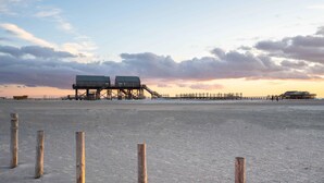 A beach with wooden posts in the foreground, a pier with buildings in the background, and a sunset sky. - Vacation home Hus rode Dör for up to 5 people (Oldenswort)