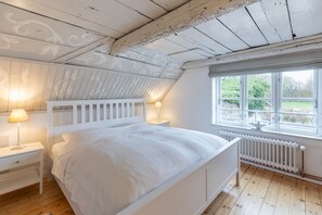 Un lit blanc avec une tête de lit, deux tables de chevet avec des lampes, une fenêtre donnant sur un paysage verdoyant et un radiateur blanc.