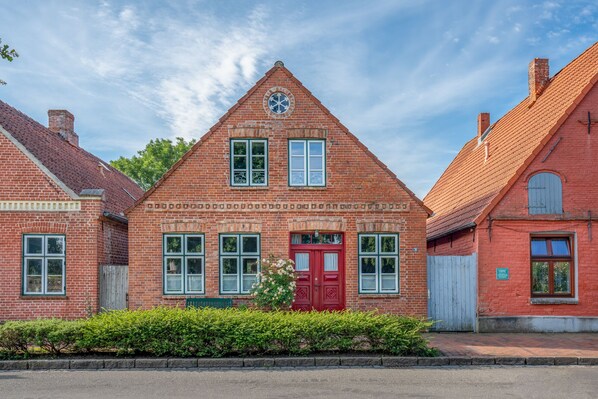 Un bâtiment en briques rouges, avec une porte rouge, de nombreuses fenêtres et un petit jardin à l’avant.
