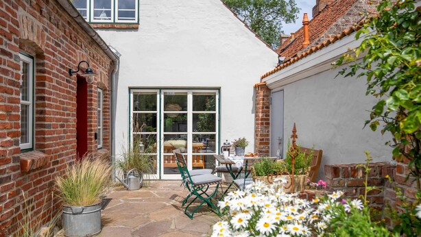 A courtyard with a white building, brick walls, a stone patio, outdoor furniture, and a variety of plants. - Vacation home Hus rode Dör for up to 5 people (Oldenswort)