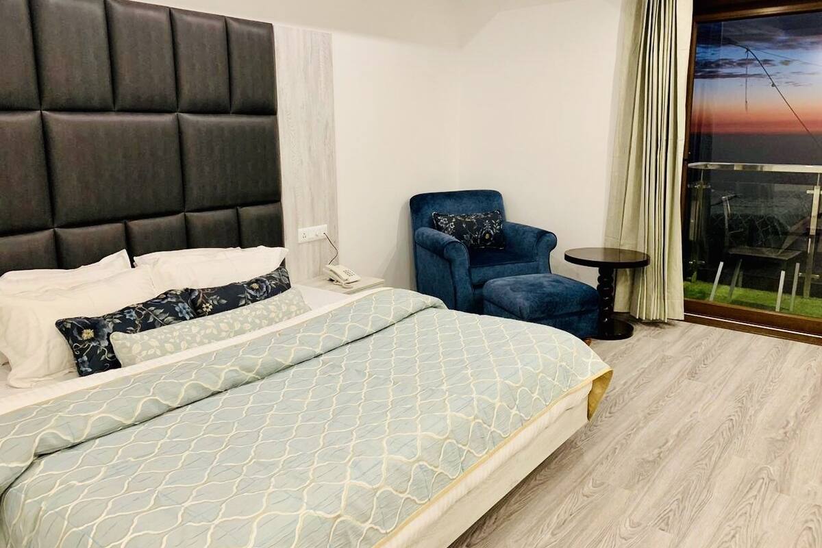 Et moderne hotelværelse med en stor seng, en blå lænestol, et lille bord og udsigt til solnedgangen gennem vinduet.