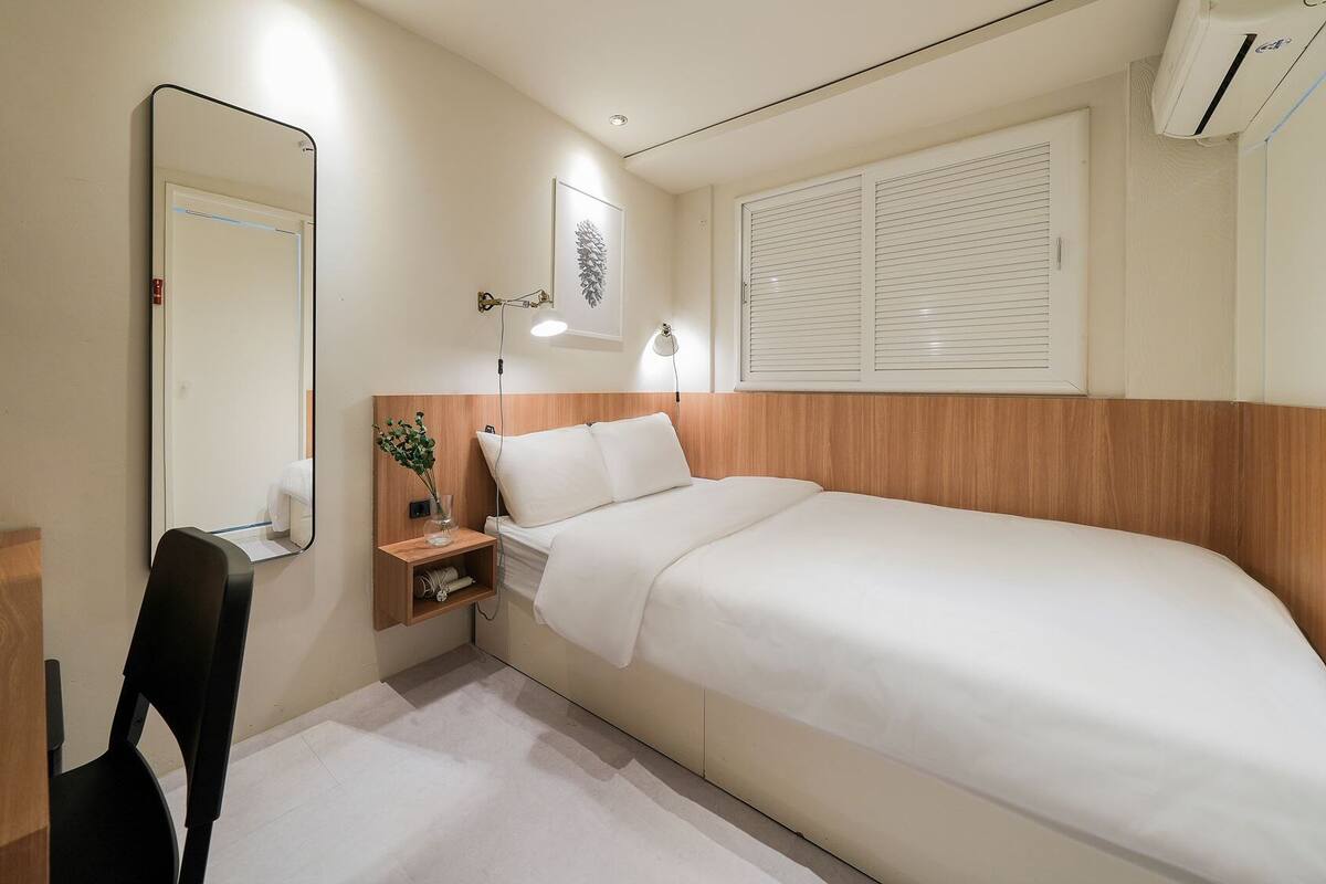 Habitación de hotel con cama, mesita de noche con lámpara, espejo y ventana con persianas.