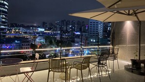 Un balcón con vistas a un paisaje urbano nocturno, que incluye una mesa y sillas bajo una sombrilla.