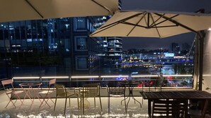 Una terraza al aire libre con mesas y sillas, un paraguas grande y vistas a un paisaje urbano nocturno.