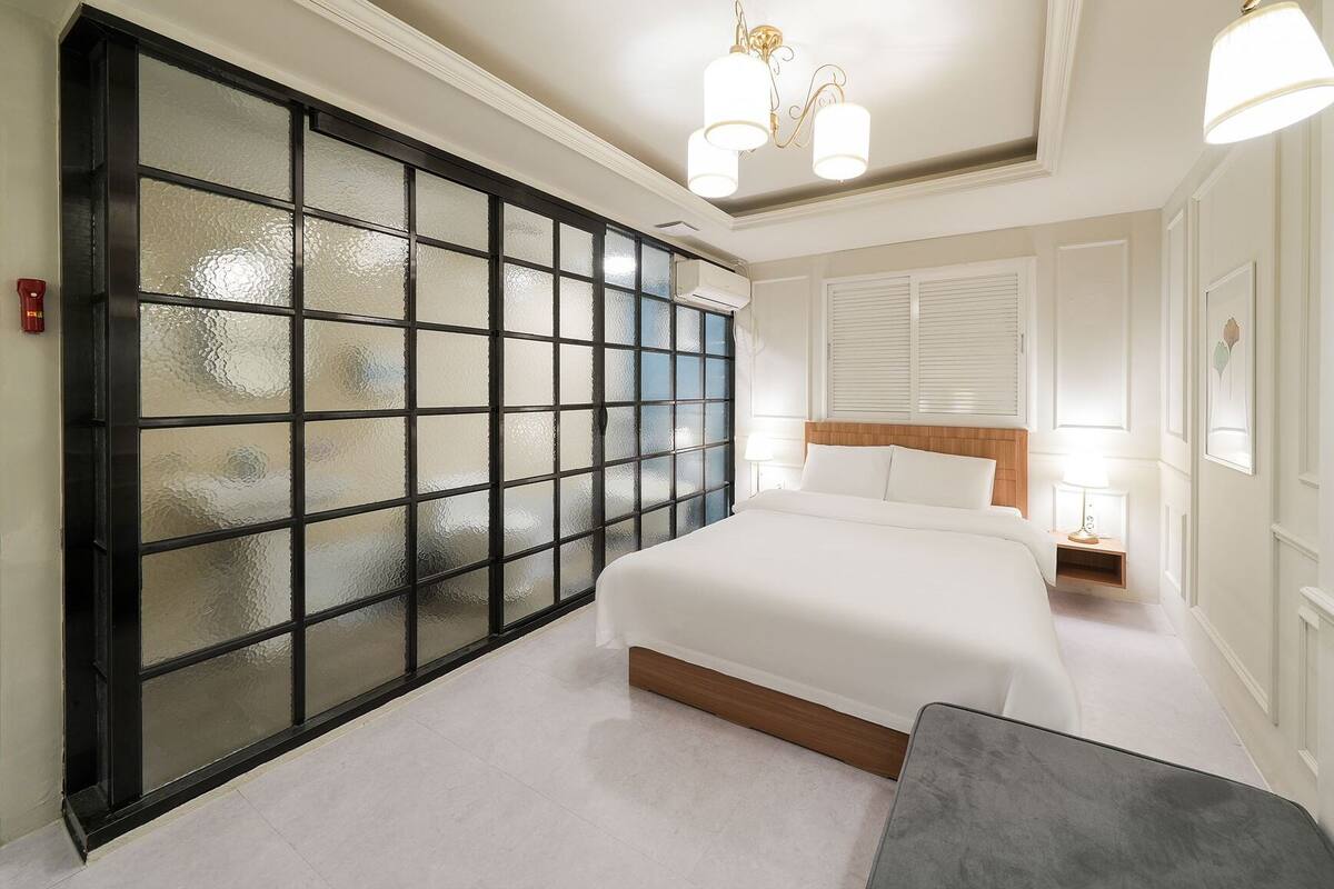Habitación de hotel con una puerta grande de vidrio esmerilado, cama con ropa blanca, mesita de noche con lámpara y lámpara moderna sobre la cama.