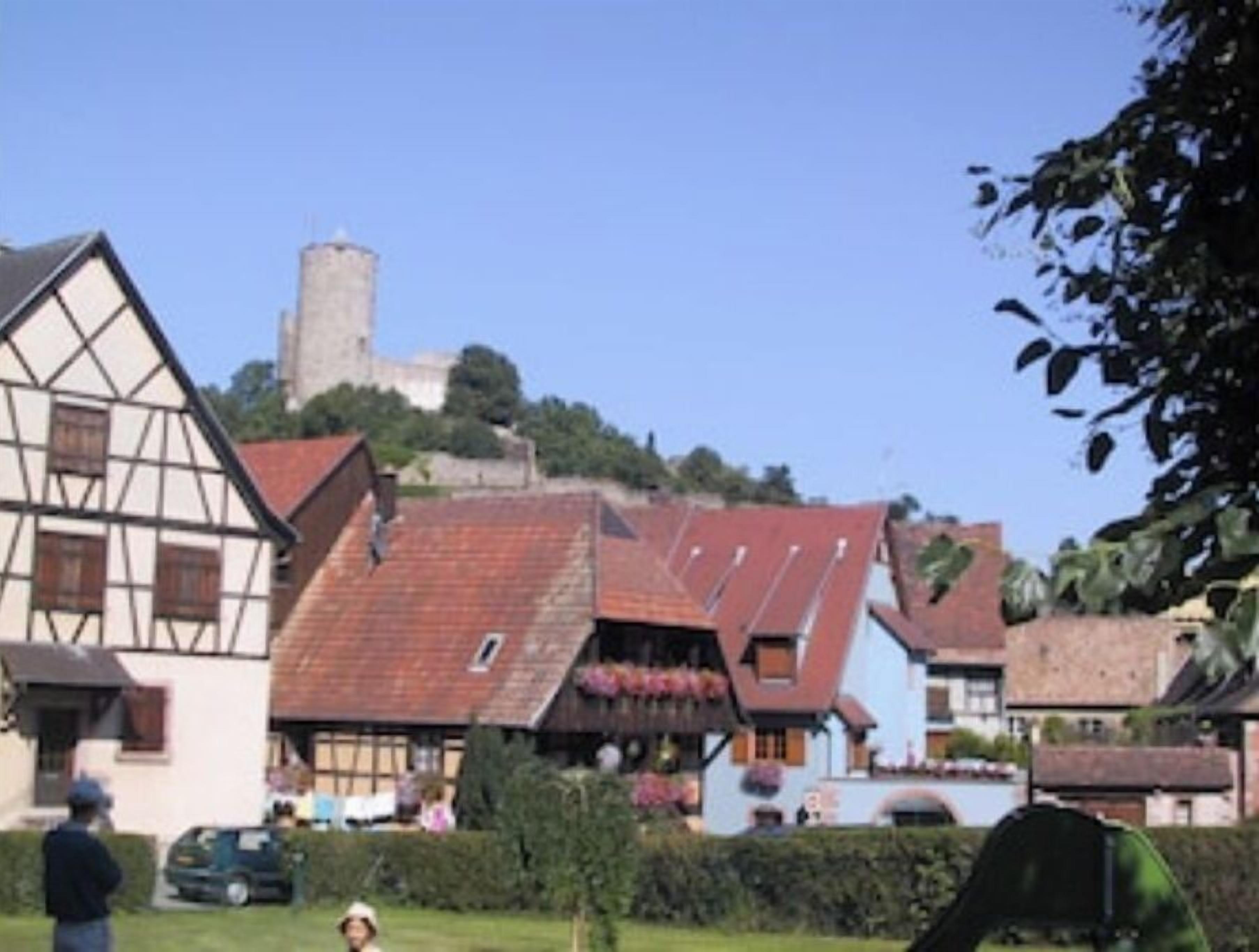 A traditional half-timbered building with a steeply pitched roof, a prominent tower, and a view of a hill with a castle-like structure in the background.
