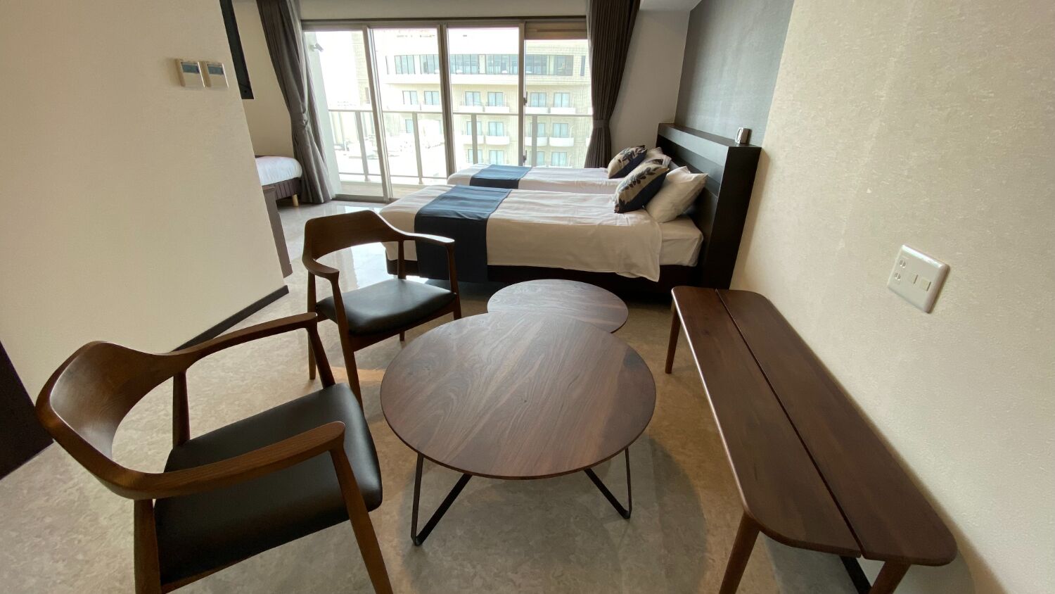 A hotel room with a bed, two chairs, a small round table, and a view of buildings through large windows.