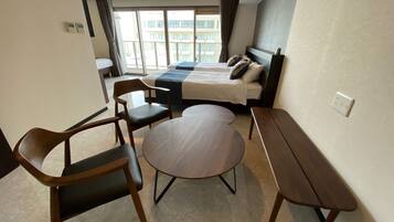 A hotel room with a bed, two chairs, a small round table, and a view of buildings through large windows.