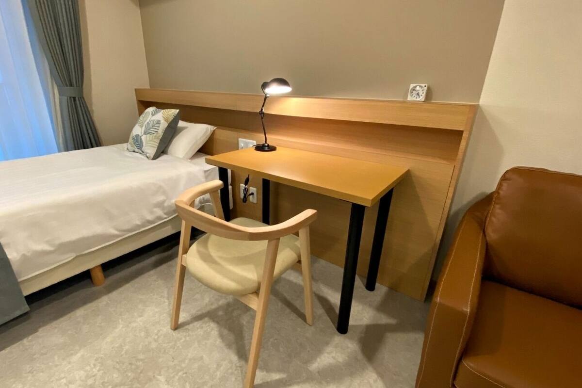 Une chambre d’hôtel avec un lit, un bureau avec une chaise, une lampe et un fauteuil en cuir marron.