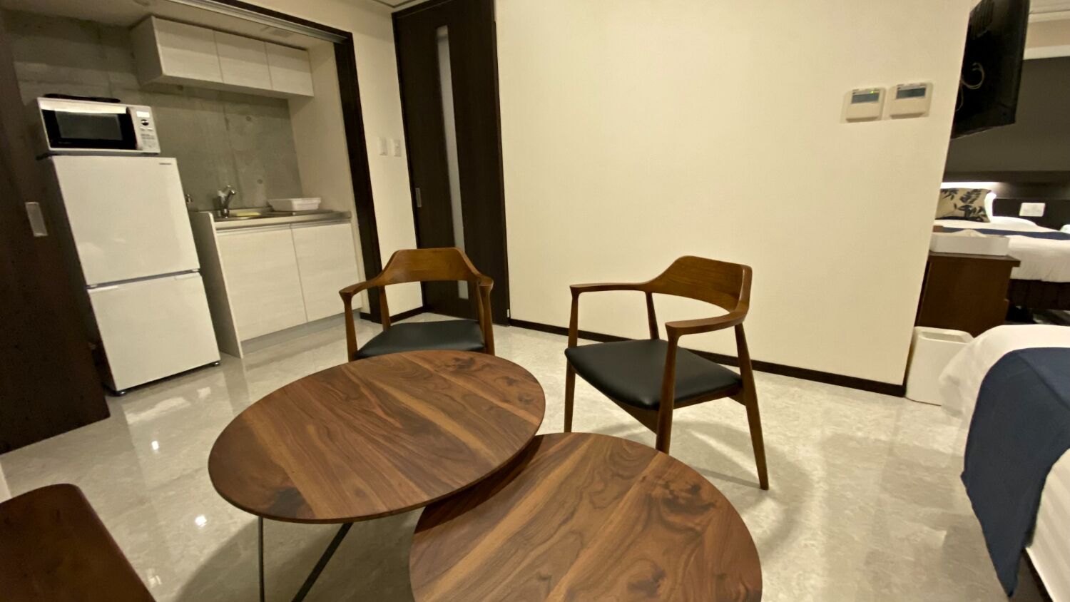 A compact kitchen with a microwave and refrigerator, two wooden chairs with black cushions, and a round wooden table.