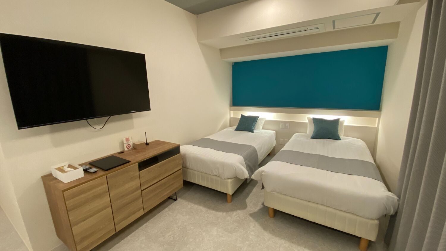 Une chambre moderne avec deux lits, un téléviseur à écran plat, une commode en bois et un mur d’accent de couleur turquoise.