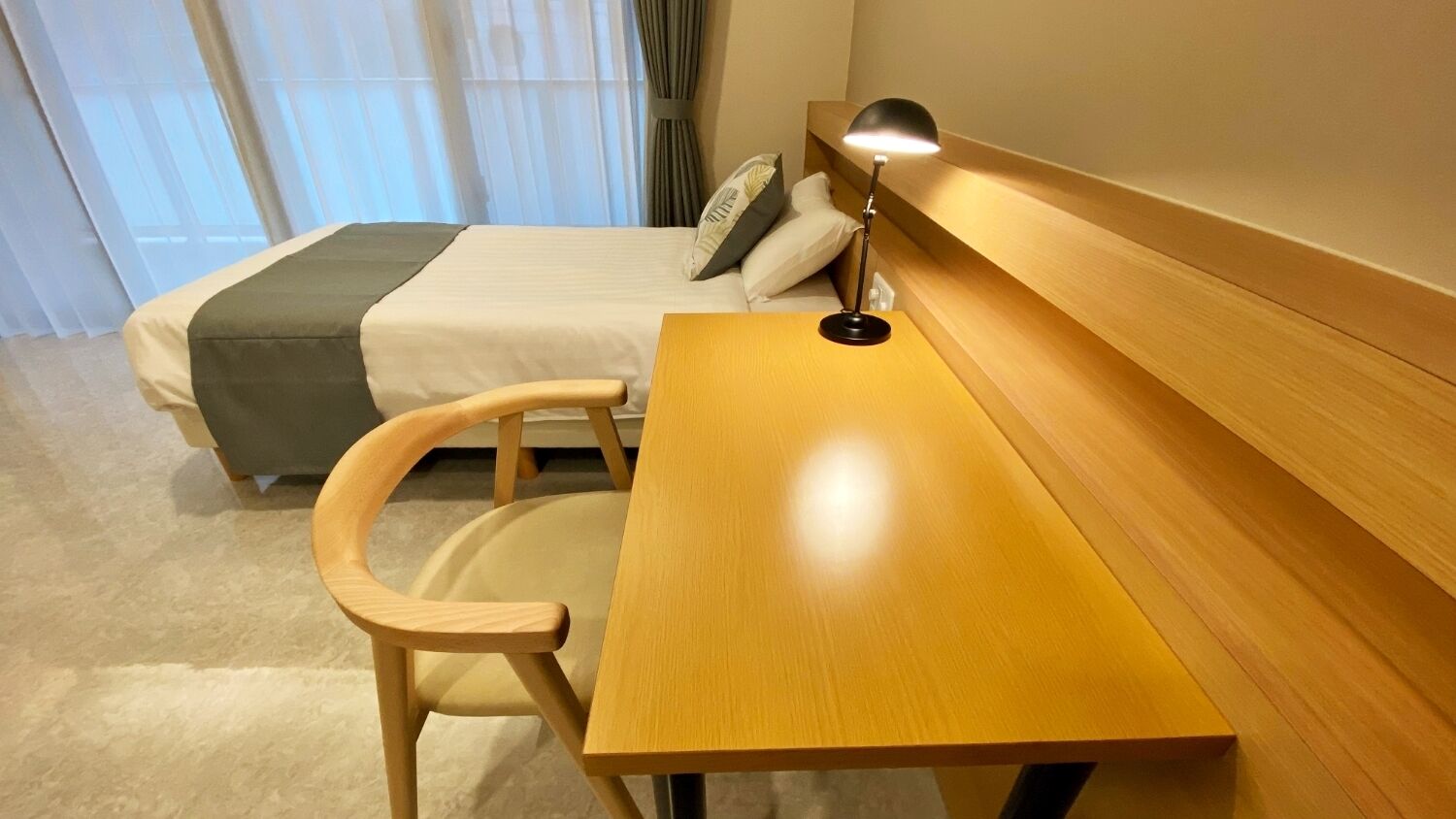 A hotel room with a bed, a wooden desk, a chair, and a lamp.