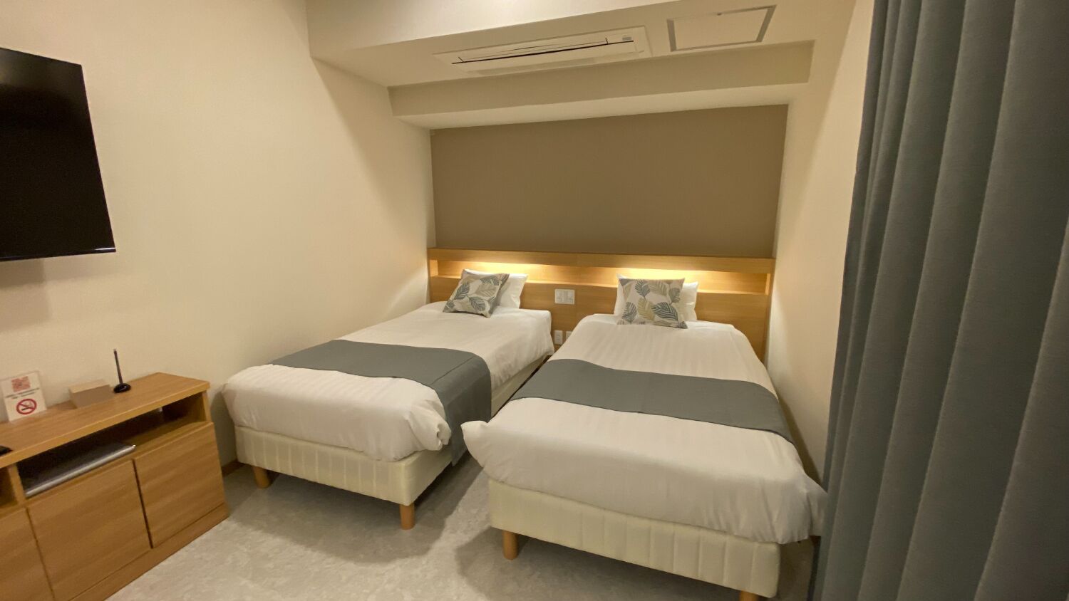 A hotel room with two beds, a wooden nightstand, a flat-screen TV, and a ceiling-mounted air conditioning unit.