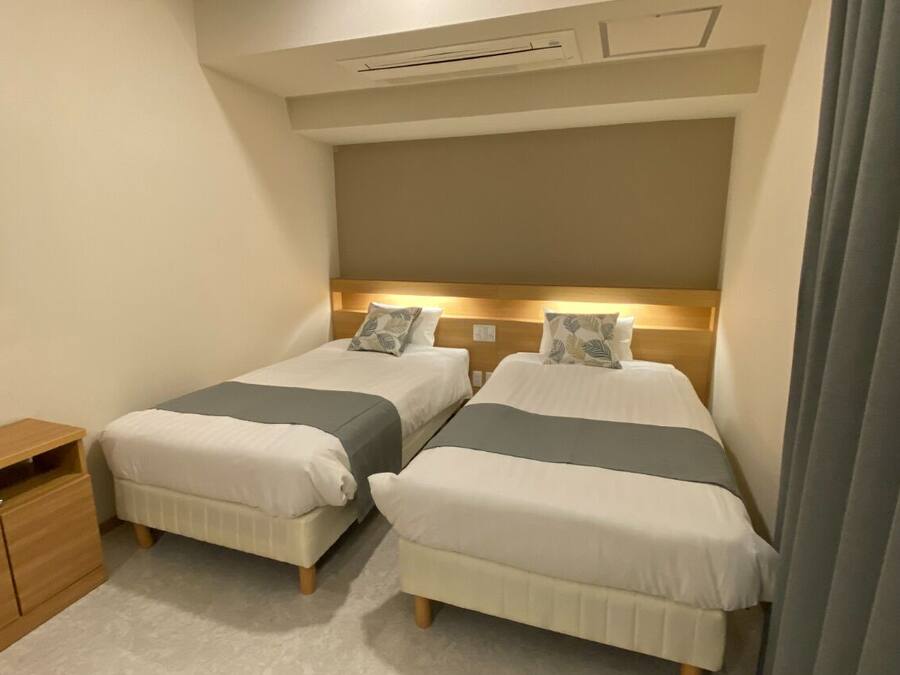 A hotel room with two beds, a wooden nightstand, a flat-screen TV, and a ceiling-mounted air conditioning unit.