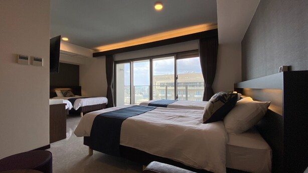 Une chambre d’hôtel moderne avec deux lits, une grande fenêtre donnant sur la ville et un coin salon séparé.