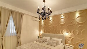 A bedroom with a large bed, a chandelier, a bedside table with a lamp, and a decorative wall panel. - Villa Vanda (Dubrovnik)