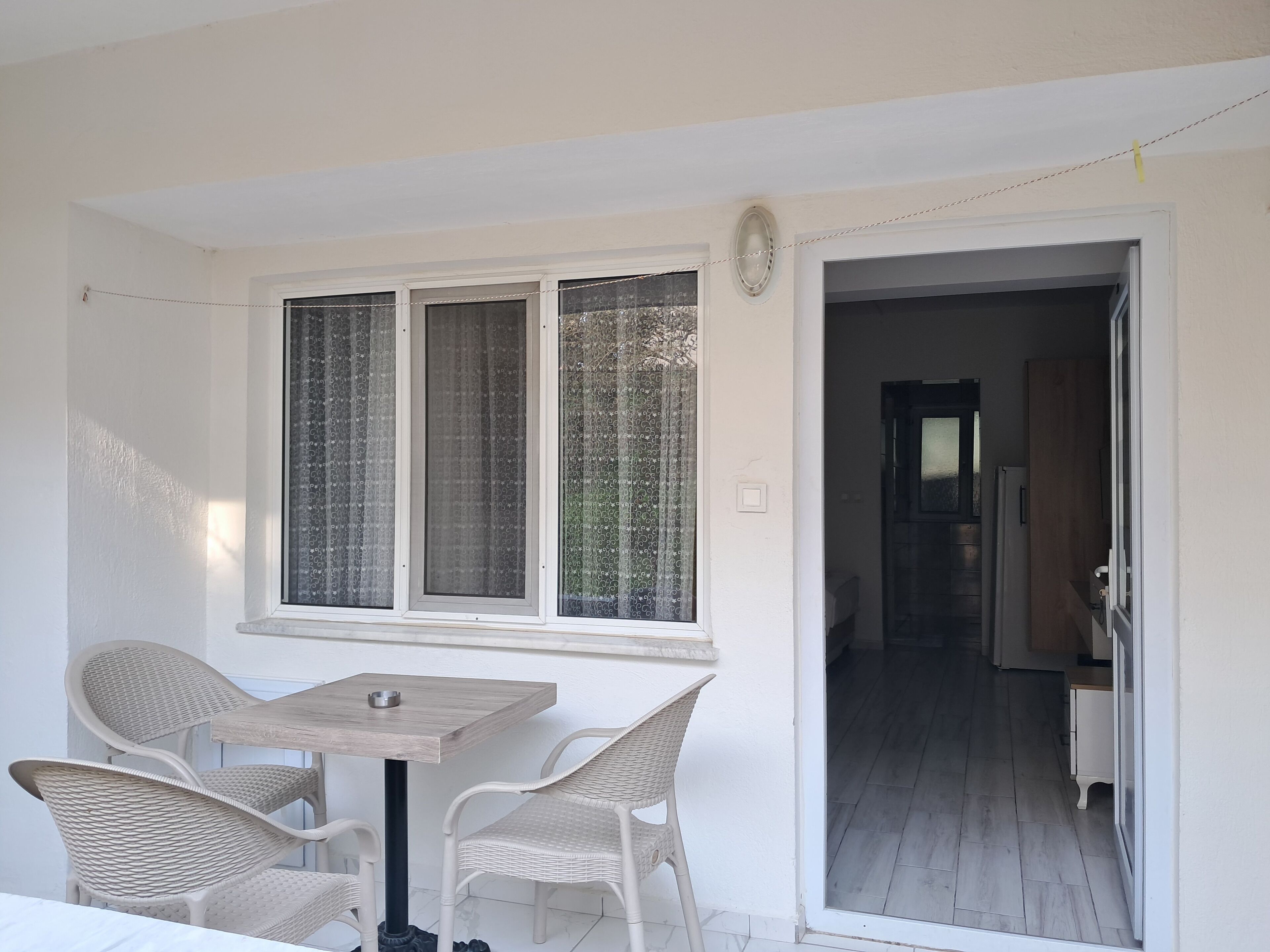 Una habitación con una mesa y cuatro sillas, un ventanal con cortinas y una puerta que da a otra habitación.