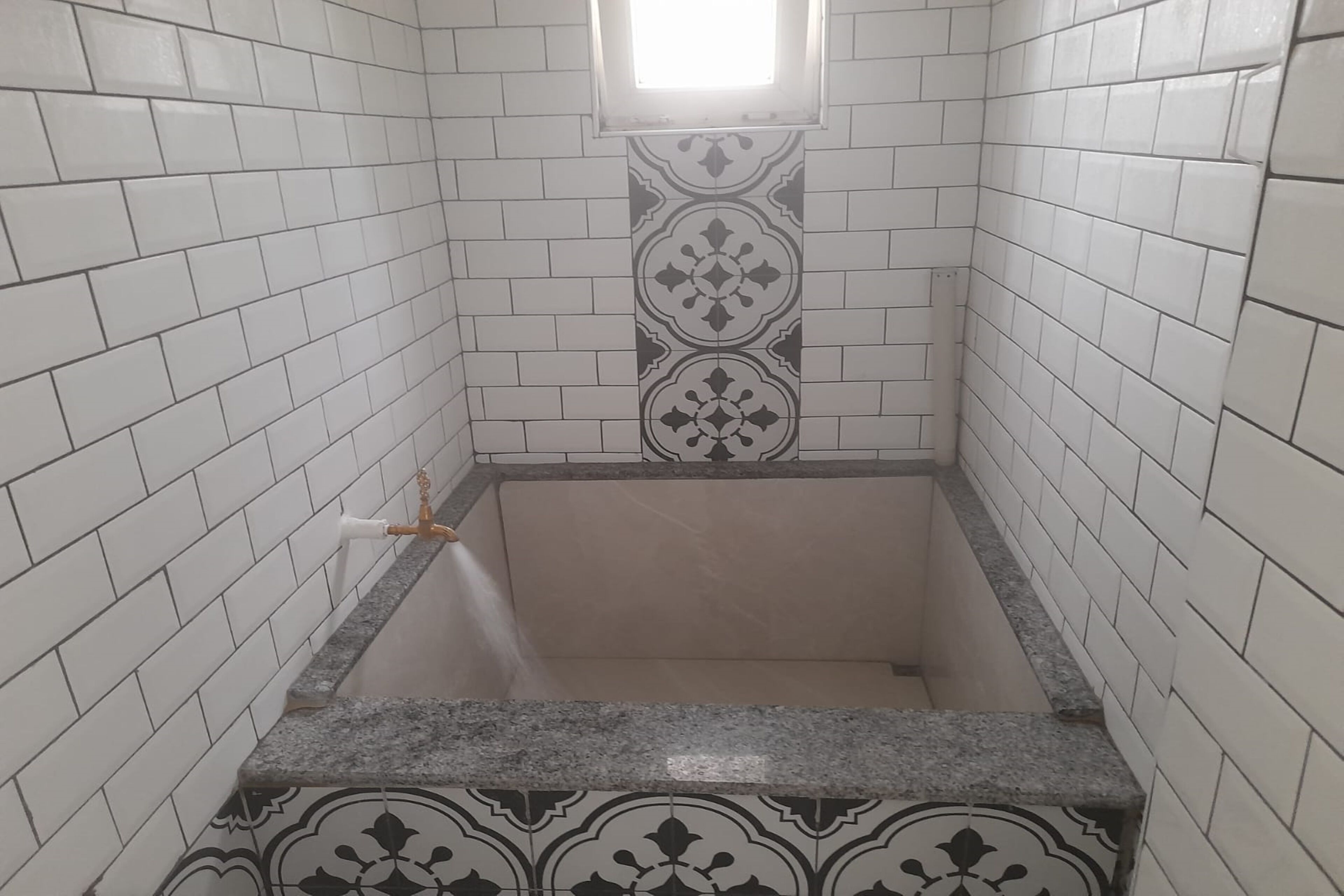A bathroom with white and patterned tiles, a sink with a faucet, and a window above the mirror.
