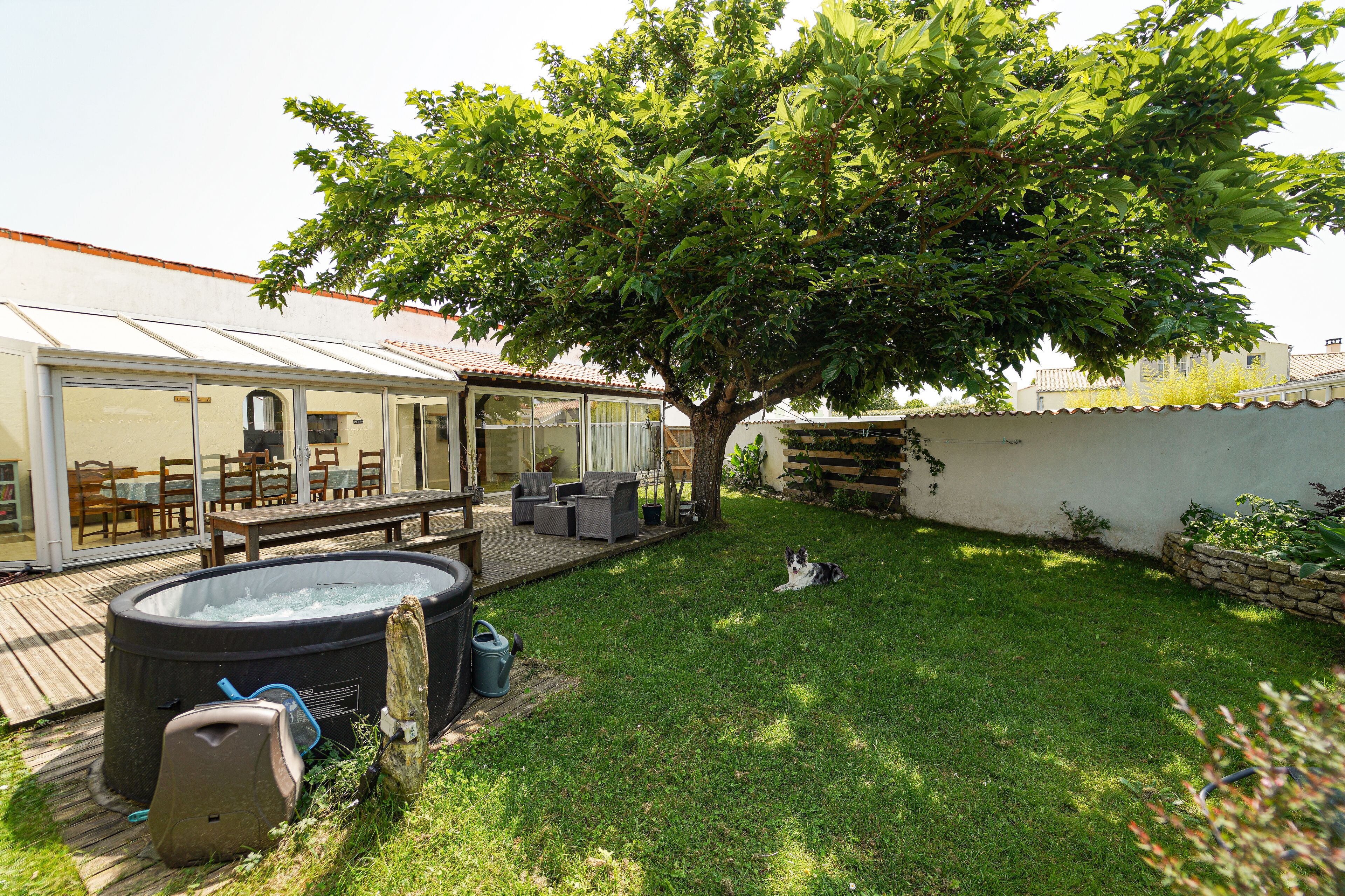 Een achtertuin met een hottub, een houten terras en een boom die schaduw geeft.