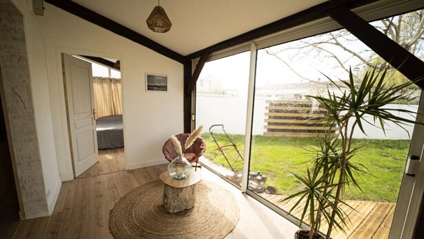 A modern room with a large window, a wooden floor, a round rug, a potted plant, and a hanging light fixture. - Oleron Surf House (Dolus-d'Oléron)