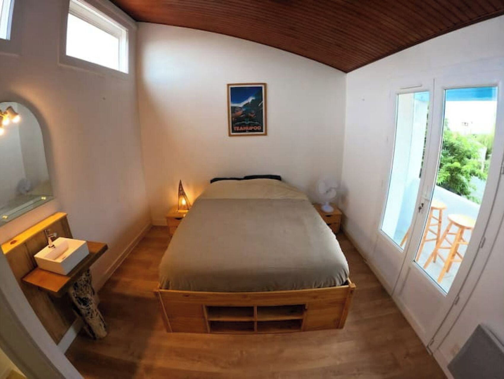 Quarto com cama de madeira, mesa de cabeceira com luminária, janela com vista para o verde e um cartaz na parede.
