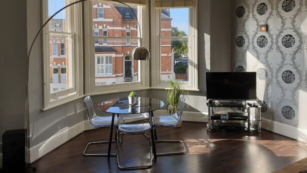 Une pièce d’angle avec une baie vitrée, une table en verre avec des chaises, une plante en pot, un téléviseur posé sur un meuble et un papier peint à motifs.