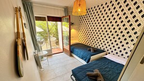 Une chambre avec deux lits, une porte-fenêtre coulissante donnant sur un balcon, un ventilateur et un mur décoré d’un motif.
