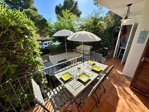 Un patio con una mesa y sillas, un paraguas blanco, y una vista de un vehículo y árboles al fondo.