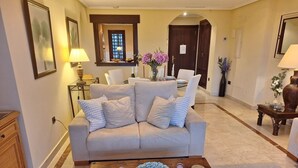 Una sala de estar con un sofá color crema, una mesa de centro de madera con objetos decorativos, y un comedor al fondo.