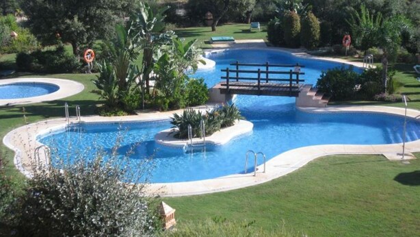 Una amplia piscina exterior con varias secciones, rodeada de césped y árboles bien cuidados.