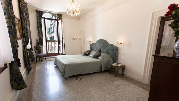 A bedroom with a bed, a chair, a nightstand, a chandelier, and a view of the street through the window. - Ca' Nal Apartments, Corte Grimani Inn - BRAGOZZO (Venezia)