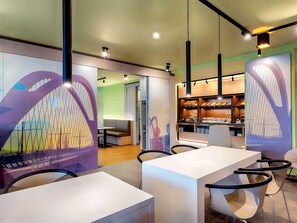 Una cafeteria moderna amb taules i cadires blanques, un separador de vidre amb un mural colorit i una barra amb exposició de menjar.