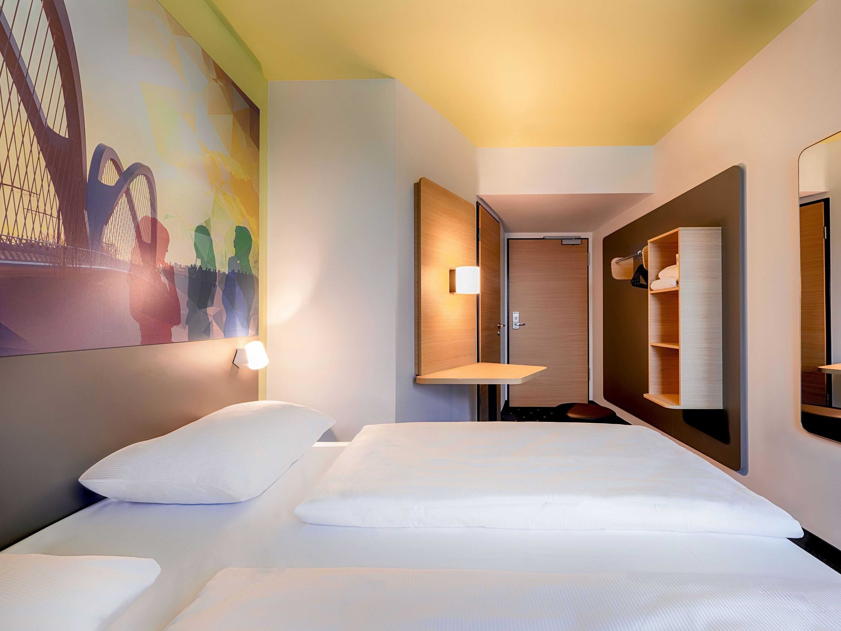 Habitación de hotel con una cama, estante en la pared, una puerta y un espejo.