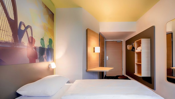 A hotel room with a bed, a wall-mounted shelf, a door, and a mirror. - B&B Hotel Kehl (Kehl)