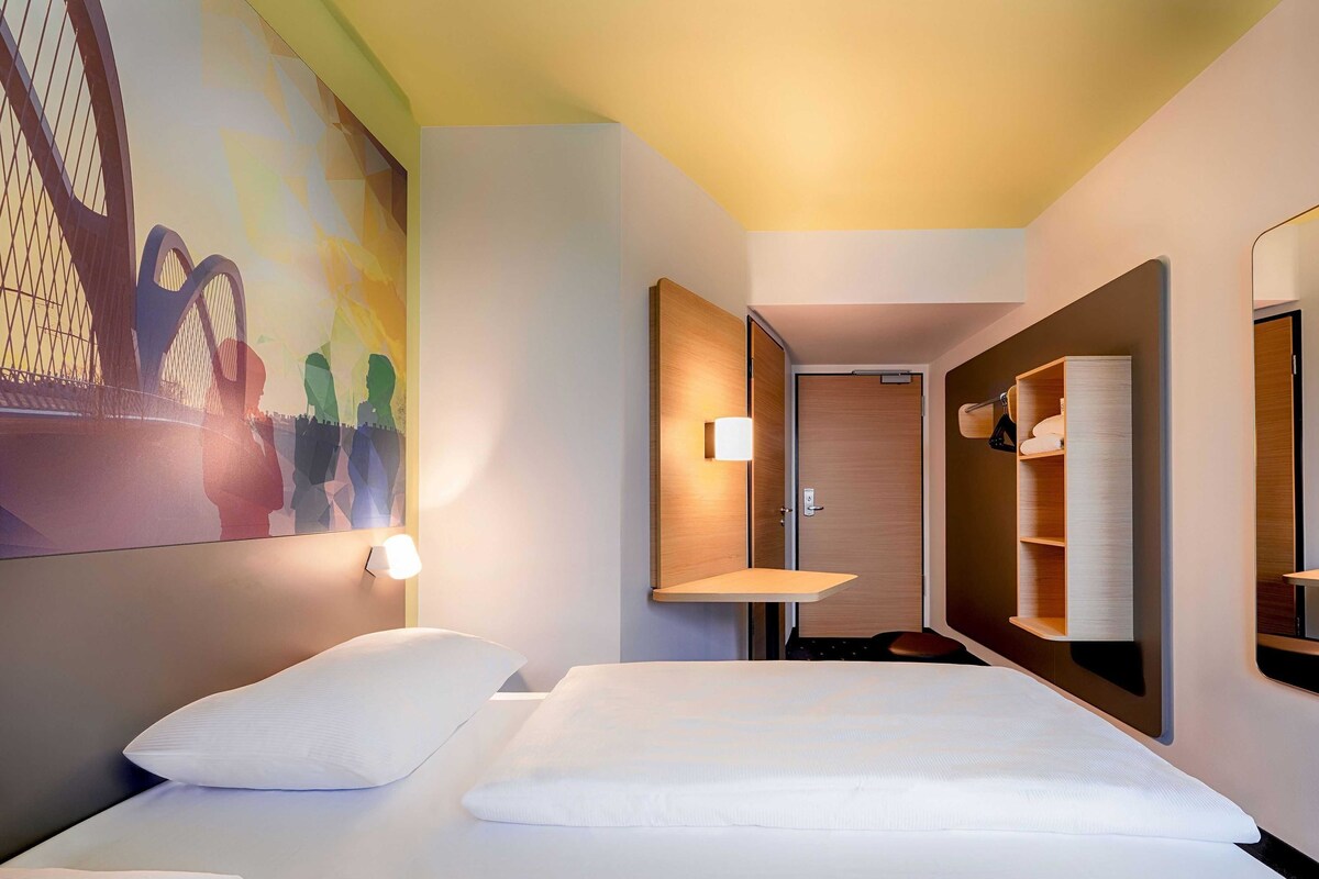 A hotel room with a bed, a wall-mounted shelf, a door, and a mirror.