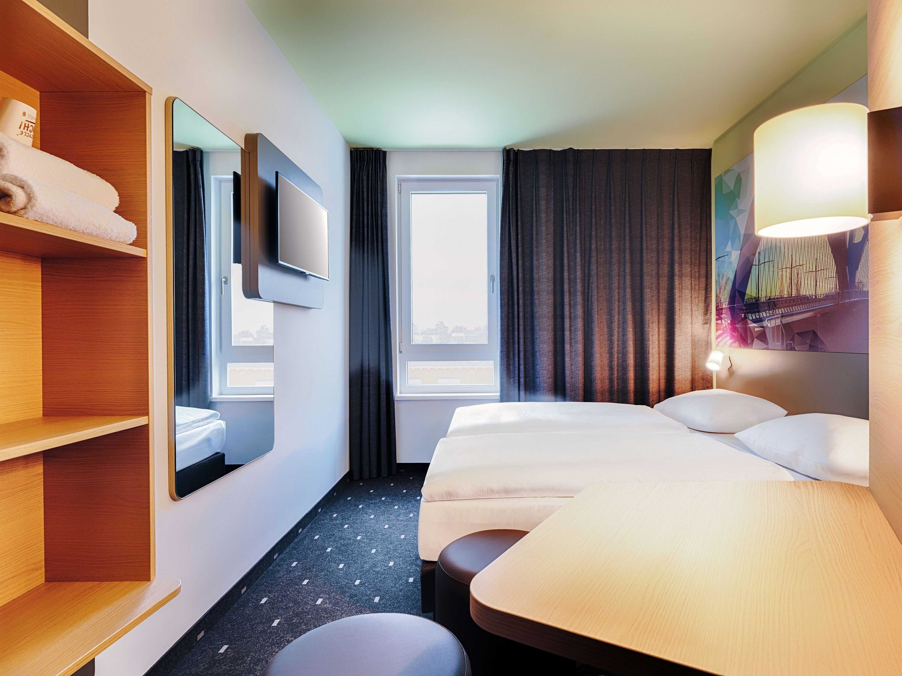 A hotel room with a large bed, a bedside table, a wardrobe, a mirror, a TV, and a window with curtains.