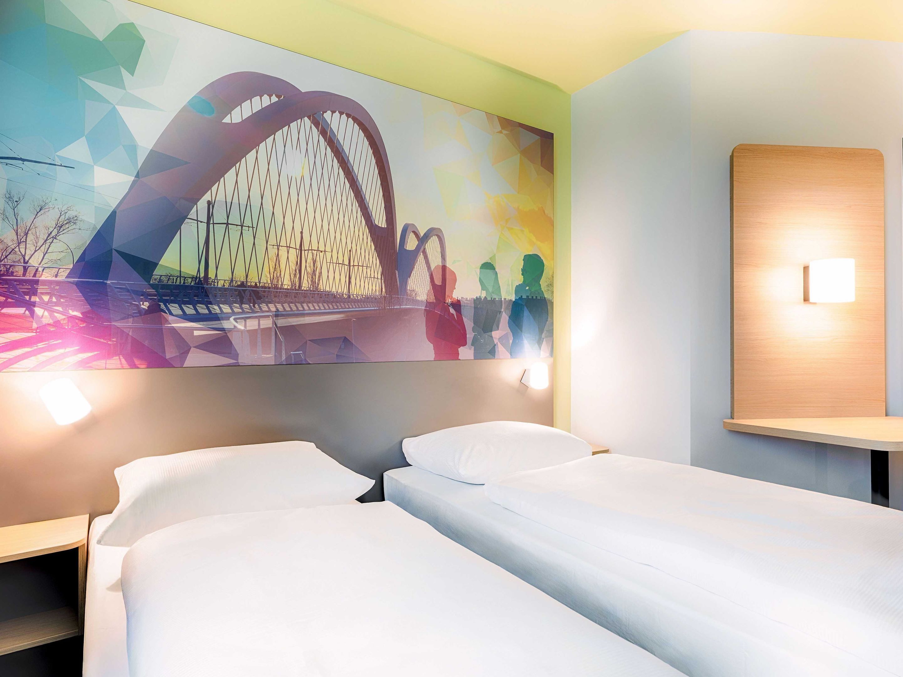 Habitación de hotel con dos camas, un gran mural abstracto en la pared y una mesita de noche con lámpara.