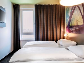 Una habitación de hotel con cama, ventana con cortinas, televisor montado en la pared y lámpara.