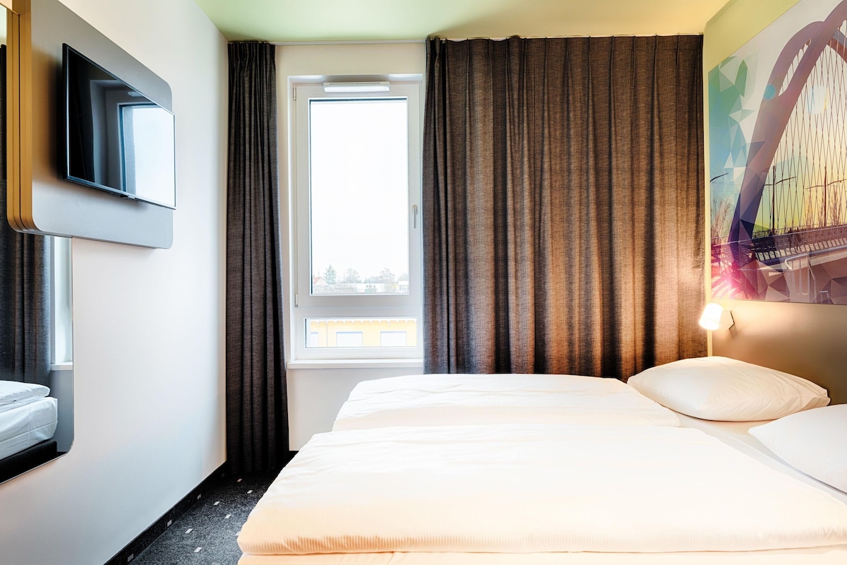 A hotel room with a bed, a TV mounted on the wall, a window with curtains, and a painting on the wall.