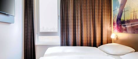 A hotel room with a bed, a window with curtains, a flat-screen TV, and a wall-mounted lamp.