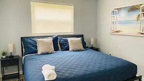 Un dormitorio con cama, ventana con persianas, mesita de noche, lámpara y un cuadro de una playa.