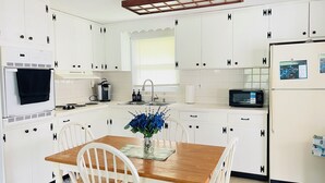 Una cocina con armarios blancos, una mesa de madera y sillas. Hay un microondas, una cafetera y un lavabo.