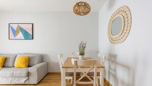 Ein modernes Wohnzimmer mit grauem Sofa, gelben Kissen, einem Holzesstisch, weißen Stühlen, einer gewobenen Leuchte und einem dekorativen Spiegel an der Wand.